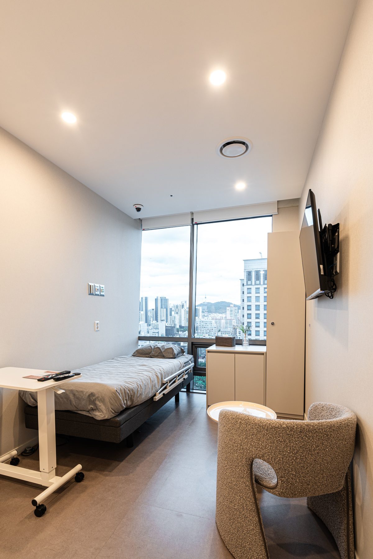 Recovery room with a hospital bed, an armchair, and a floor-to-ceiling city view
