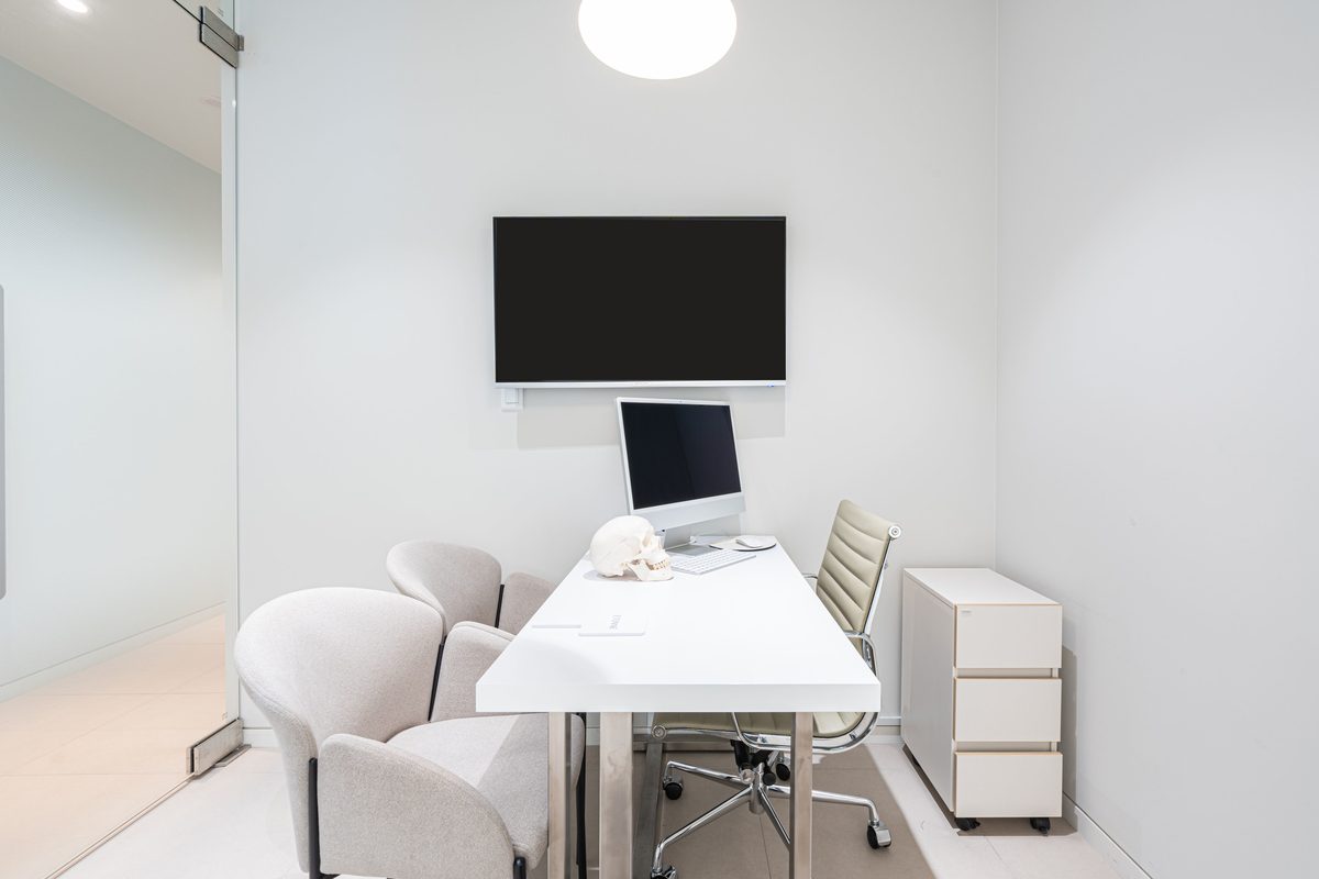 Consultation room with a wall-mounted screen, desk, and skull model seen through a glass door